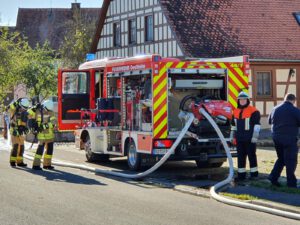 der Maschinist steht hinter dem Feuerwehrfahrzeug und bedient die Pumpe, während sich ein Trupp neben dem Fahrzeug mit Atemschutz ausrüstet.